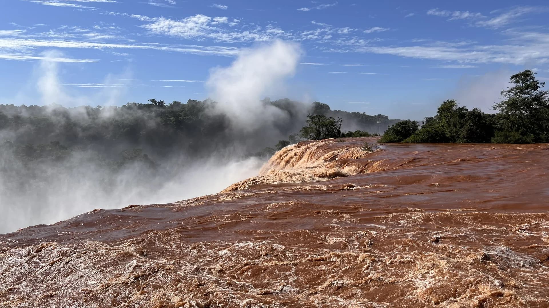 The Iguaçu River in flood at the brink of the falls, running red with sediment, with spray rising into the forest on the far bank