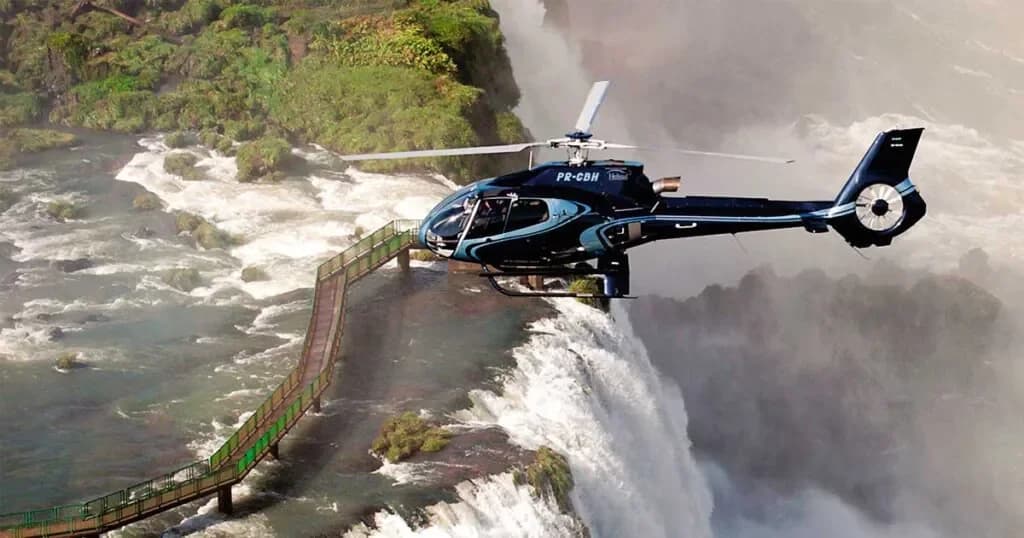 Helicopter registered PR-CBH banking over Iguazu Falls, with a park walkway on the rock ledge below