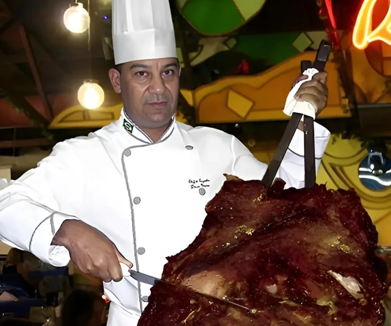 A chef in whites slicing from a large cut of meat at a carving station at Rafain Churrascaria in Foz do Iguaçu, under the restaurant's stained-glass ceiling